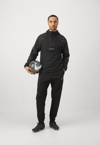 Zwarte Nike trainingspak met een capuchonjack en tapered broeken. Weergegeven met een gestructureerde voetbal. Nike logo's op de kleding en de bal.