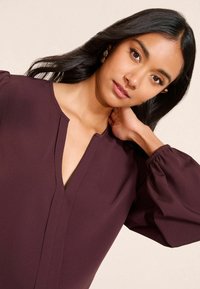 Burgundijas blūze ar V veida dekoltē, garām pūkainām piedurknēm ar elastīgām aprocēm un gludu, vieglu auduma tekstūru.