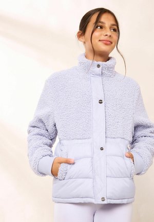Young woman wearing a light purple sherpa and quilted jacket with hands in pockets, looking to the side with a slight smile.