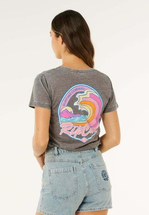 Femme aux longs cheveux bruns portant un t-shirt gris de la marque de surf Rip Curl et un short en denim clair, debout dos à la caméra.