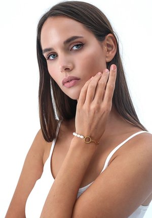 Zlatý náramok s bielymi perlovými akcentmi a kruhovým zdobením. Modelova ruka jemne spočíva na jej tvári, čo zdôrazňuje dizajn šperku.