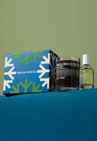 Schwarze Kerze und hellgrüne Ölflasche mit Etikett, verpackt in einer blauen Schachtel mit weißen und grünen Schneeflockenmuster.