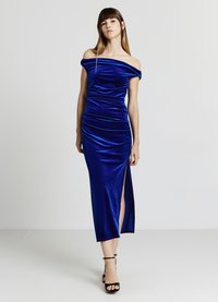 Blaues Samtkleid mit Carmenausschnitt, figurbetontem Oberteil, seitlichen Raffungen und hohem Beinschlitz. Gestylt mit schwarzen Absatzschuhen mit Knöchelriemen.