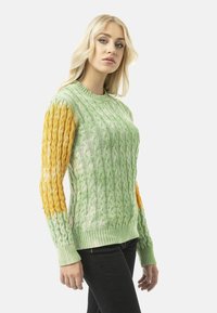 Groen en gele kabelgebreide trui met een textuur patroon, aangesloten silhouet, geribde manchetten en ronde halslijn, gedragen met zwarte broek.
