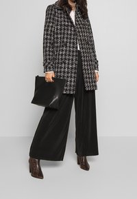 Svart och vit rutad kappa med avslappnad passform, bärs över en ljus skjorta, i kombination med vida svarta plisserade byxor och bruna klackstövlar.