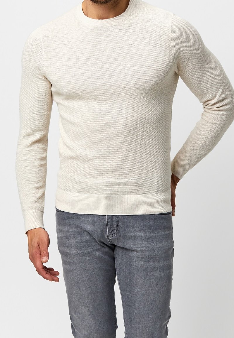 Homme portant un pull en maille texturée beige clair, ajusté, et un jean gris délavé, debout avec une main dans le dos.