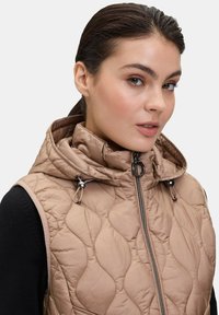 Gilet beige trapuntato con colletto alto e cappuccio, caratterizzato da dettagli in zip neri e lacci, realizzato in un materiale liscio e lucido.