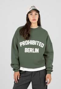 Moteris vilkinti žalią švarką su užrašu "PROHIBITED BERLIN", pilkas kelnes, baltą marškinėlį, smėlio spalvos kepurę su suderintu užrašu "Prohibited 10119 Berlin".