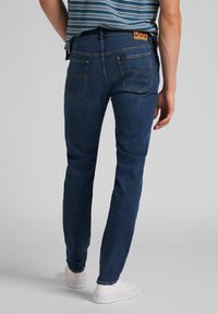 Blå denimjeans med slim fit, som har två bakfickor och en logotyppatch i brunt läder. Tyget har en slät textur.