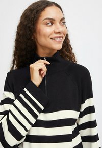 Pull noir et blanc à rayures avec col haut et détails zippés, en tissu doux et avec des manches larges.
