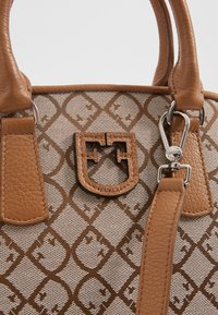 Sac à main à motifs marron clair et beige avec poignées en cuir, écusson central en cuir avec logo et fermoir argenté sur une sangle en cuir.