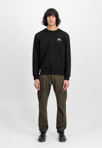 Svart hettegenser med rund hals og logo på brystet, kombinert med olivengrønne cargo-bukser med lommer. Modellen har på seg sorte sko.