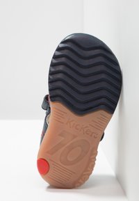 Suela de zapatilla de goma negra con textura ondulada. Presenta un acento rojo y el nombre de la marca grabado, con un borde exterior beige que muestra la talla "10".