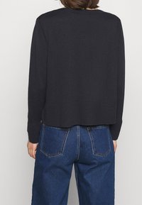 Osoba ubrana w czarny sweter z długim rękawem i niebieskie jeansy z wysokim stanem, pokazana od tyłu na jednolitym tle.