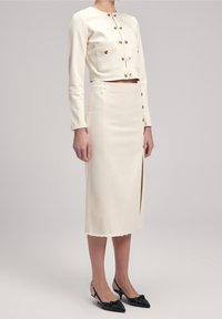 Chaqueta de mezclilla crema con botones dorados y dos bolsillos delanteros, combinada con una falda lápiz a juego con una abertura frontal y dobladillo desflecado. Zapatos negros tipo slingback.