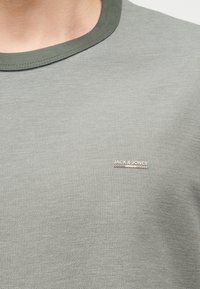 Κλείσιμο πλάνου ενός ατόμου που φοράει ένα ανοιχτό γκρι υφασμάτινο μπλουζάκι με σκούρο πράσινο ριμπ λαιμόκοψη και το λογότυπο "JACK & JONES" στο στήθος.