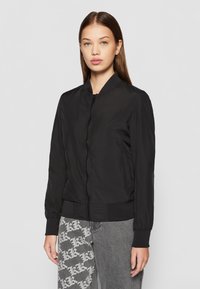 Crni bomber jakna s patentnim zatvaračem sprijeda, rebrastim manžetama i elegantnim dizajnom. Izrađena od glatkog, laganog materijala.