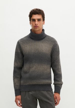 Jeune homme portant un pull à col roulé côtelé gris et beige et un pantalon foncé, debout devant un fond clair uni.