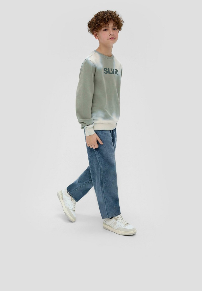 Lichtgroene tie-dye sweatshirt met zwarte "SLVR" tekst, gecombineerd met loszittende blauwe denim broek en witte sneakers. Casual en ontspannen pasvorm.