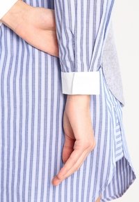 Camisa de rayas verticales azul y blanca con puños blancos. Cuenta con una tela suave y aberturas laterales para mayor comodidad de movimiento. La parte trasera es de color gris.