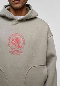 Beige hoodie met een grote ronde rode grafiek met een roos en tekst. Het heeft een voorzak en een capuchon met trekkoord. Zachte stof textuur.