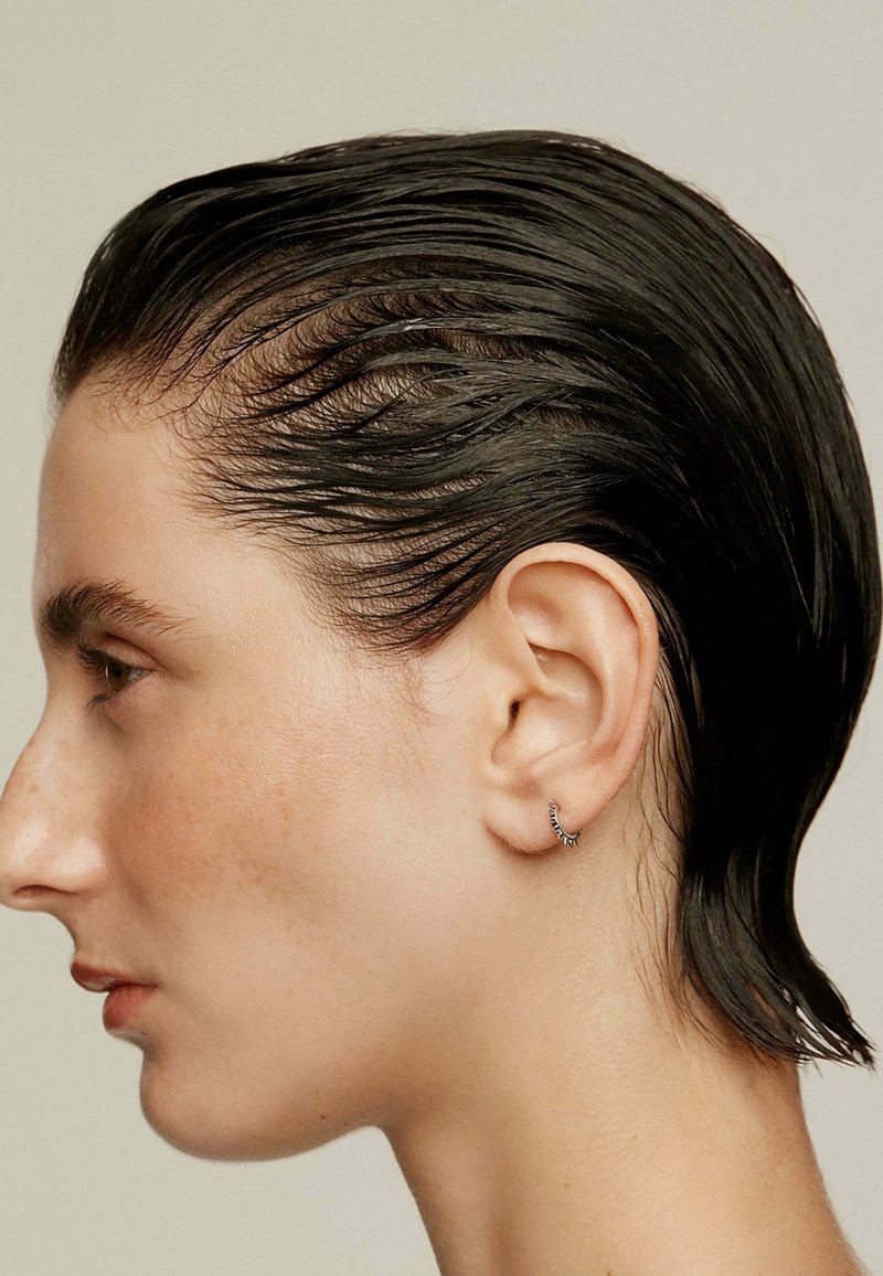 Vista lateral de una persona con cabello oscuro y liso, y un pequeño pendiente de aro texturizado con sutiles adornos. Piel suave y rasgos definidos.