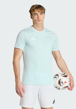 Młody mężczyzna w jasnoniebieskiej koszulce Adidas i białych szortach trzyma obok siebie białą i czarną piłkę nożną Adidas.