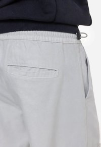 Gros plan sur un pantalon gris clair à taille élastique avec une poche passepoilée au dos et un cordon de serrage noir réglable sous un haut bleu marine foncé.