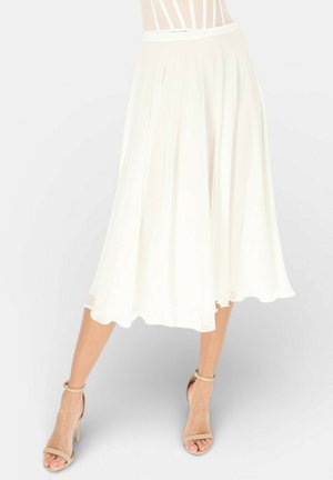 Leciutka biała spódnica midi noszona z beżowymi wysokimi obcasami z paskiem wokół kostki, nogi lekko rozstawione na jasnym, jednolitym tle.