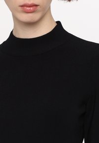Svart höghalshöjd topp i ribbat material, figursydd design, långa ärmar, med en mjuk textur, som visar en del av halsen och nedre ansiktet.