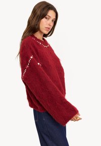 Pull en laine rouge duveteux avec un col rond, présentant des accents argentés le long des épaules et des manches. Manches larges et amples en tissu texturé.