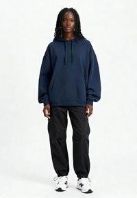 Felpa oversize blu navy con tasca frontale e cappuccio regolabile, abbinata a pantaloni cargo neri affusolati e sneaker bianche.