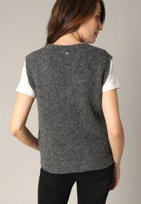 Gilet gris en tricot avec une texture côtelée, des manches courtes et un col rond. Le dos présente un petit détail de bouton. Porté sur une chemise blanche.