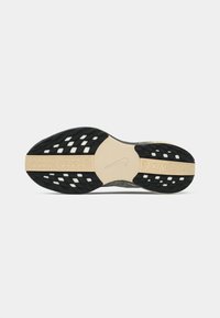 Sål af en Nike løbesko med sorte teksturerede greb og en beige central stripe med Nike-logoet og tekst.