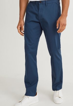 Muž oblečený v tmavě modrých kalhotách s rovnými nohavicemi a bílých teniskách, stojící před jednobarevným světlým pozadím.