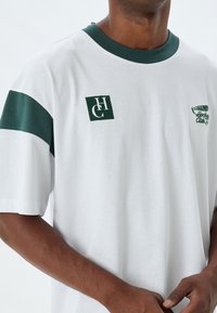 Camiseta de algodón blanca con acentos verdes. Presenta un cuello redondo verde y mangas rayadas. En el pecho izquierdo tiene logotipos y detalles bordados en verde.