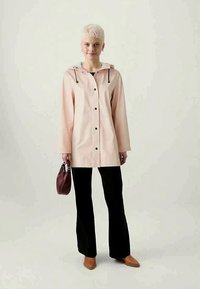 Chaqueta impermeable de color rosa claro con capucha, con cordones y botones negros. Llevada sobre pantalones negros y combinada con un pequeño bolso oscuro.