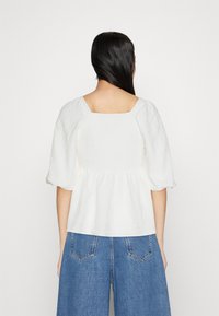 Blusa bianca con maniche a sbuffo, scollo quadrato e tessuto texturizzato, caratterizzata da una vita arricciata e una silhouette fluida.