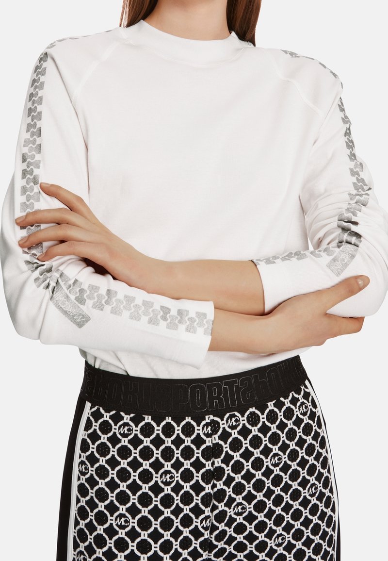 Hvid sweatshirt med raglan-ærmer og grå mønstrede detaljer langs armene, kombineret med sorte mønstrede træningsbukser.