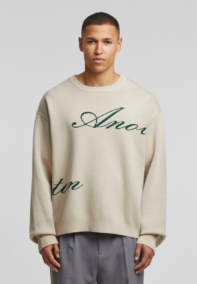 Beige gebreide trui met een oversized pasvorm, voorzien van groene cursieve tekst. Ribbelboord bij de hals en mouwen. Soepele textuur, casual ontwerp.