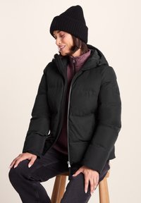 Woman wearing black beanie, black padded jacket, and purple zip-up sweater, sitting on wooden stool against beige background.