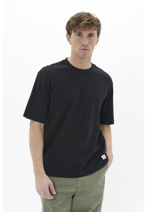 T-shirt nero a maniche corte in tessuto a coste, con scollo rotondo e una piccola etichetta sull'orlo laterale. Abbinato a pantaloni verde oliva.