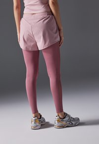 Shorts de sport roses clairs superposés sur des leggings roses assortis, avec une texture lisse et une taille élastique. Des chaussures de sport argentées et dorées complètent la tenue.