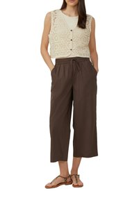 Häkelweste in Creme mit hexagonalen Mustern, über einem beigen Oberteil getragen. Braune, locker sitzende Hose mit elastischem Bund und Seitentaschen.