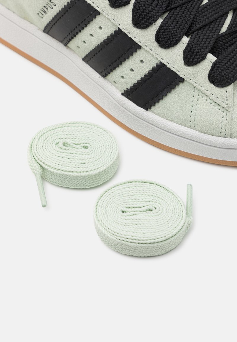 Sneaker verde in suede con strisce nere, caratterizzato da una suola bianca e lacci rotondi verde menta posizionati accanto alla scarpa.