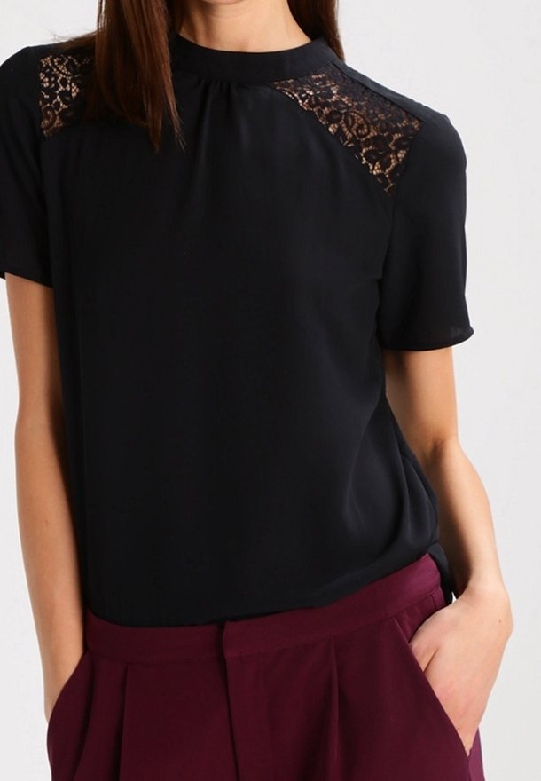Blusa negra con mangas cortas, que presenta detalles de encaje en los hombros. Está hecha de una tela suave, con un cuello alto y un corte holgado.