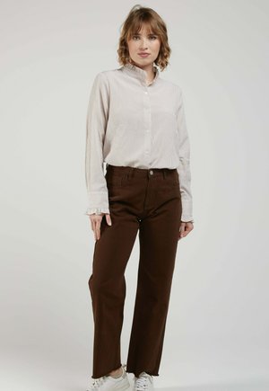 Femme portant un chemisier clair à rayures avec des poignets et un col à volants, un pantalon marron foncé et des baskets blanches, debout devant un fond uni.