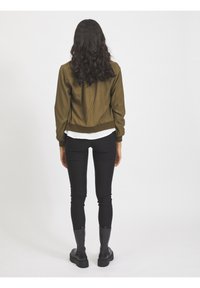 Femme aux longs cheveux bouclés portant une veste verte olive, une chemise blanche, un pantalon noir et des bottes noires, se tenant de dos sur un fond blanc.