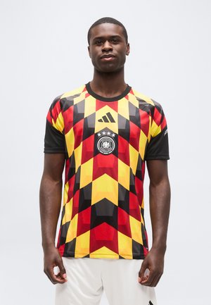 Homme portant un maillot de football d'Allemagne avec un motif géométrique rouge, jaune et noir et le logo de l'équipe, debout devant un fond blanc.