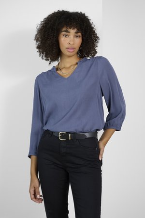V-Ausschnitt, langärmlige Bluse aus weichem blauen Stoff, kombiniert mit schwarzen Jeans und einem schwarzen Gürtel mit einer goldenen Schnalle. Schlichtes, strukturiertes Design.